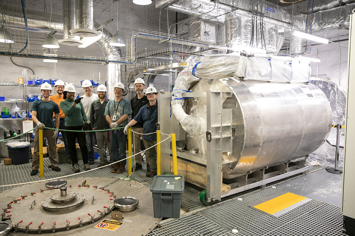 Eight people stand to the left of a large piece of the LZ dark matter detector 