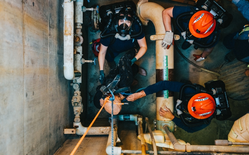 a person wearing a full-face breathing apparatus looks up through a utility hole as the emergency rescue team raises a training mannequin using ropes.