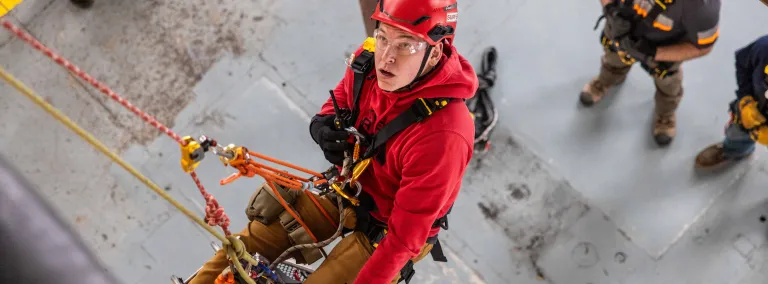 Tyler Schwindt, a hoist operator at SURF who is a also member of the ERT, trains on the new tower while lifting a medical training manikin in a stretcher.  He is wearing a hemlit and proper PPE for the job and is supported by two seperate ropes while hanging on a wall suspendede over two people who are lookiing up at him. 