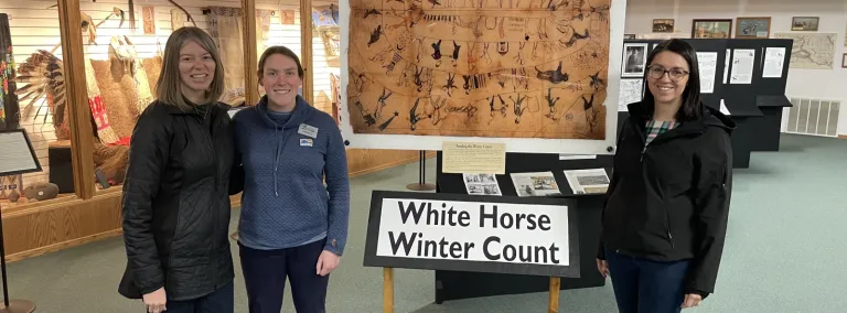 Kelly Kirk (left), Director of the Sanford Lab Homestake Visitor Center, along with Emily Berry and Rochelle Zens, who are part of the outreach and culture team at SURF, pose in front of the White Horse Winter Count at the Timber Lake Museum.