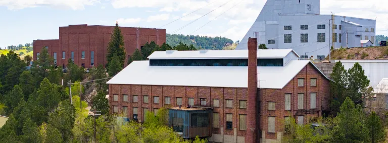 An aerial view of the Foundry Building at SURF, showing a brick building with the Yates headframe and hosstroom in the background surrounded by green trees in the spring. 