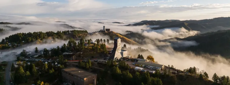 The Ross and Yates headframes, at the Sanford Underground Research Facility, sit above the clouds covering the Black Hills in October of 2023. This image of fog rolling in between the Ross and Yates headframes at the Sanford Underground Research Facility (SURF) during a sunrise in October of 2023. In rare cases, fog will cover the lower lying valleys and hills around SURF, leaving the town of Lead exposed in the sunlight, above the clouds, like an island on the ocean.