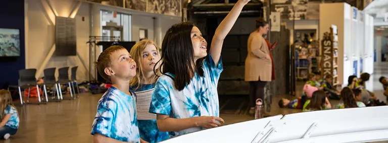 Students engage in STEM learning at the Sanford Lab Homestake Visitor Center in lead.  Three children are looking up at a model hanging from the celing. 