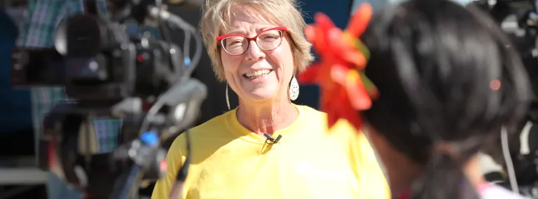 Constance Walter, wearing a bright yellow shirt and red glasses, smiles during TV interviews through a crowd of people.