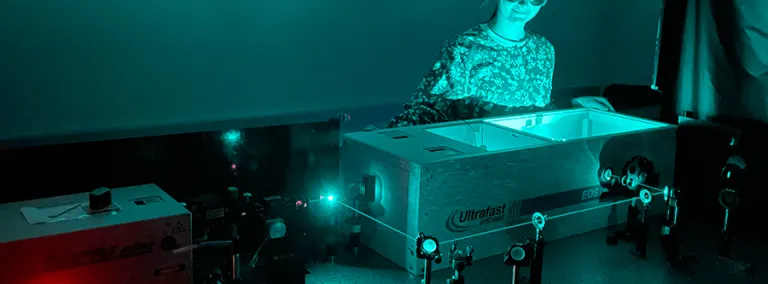 Madison Jilek works in her laboratory at the University of Colorado Boulder.  She is wearing laser protective eyeware, in a room illuminated by green light with a green colored laser running through a series of lenses and mirrors around a table. 