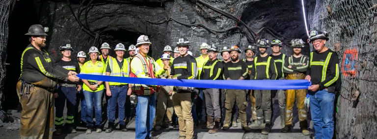 A group of poeple stands in a newly excivated drift a ribbon is being cut with large scissors, everyone is in PPE and smiling.   