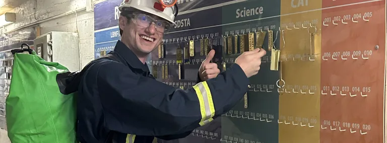 Archer Sagaskie, a student of physics at Grand Valley State University in Michigan smiles as he places a brass tag on the board before going underground at SURF.  Archer is in full SURF PPE, including a hard hat, overalls, safety glasses, and a W65 resperator on his belt.