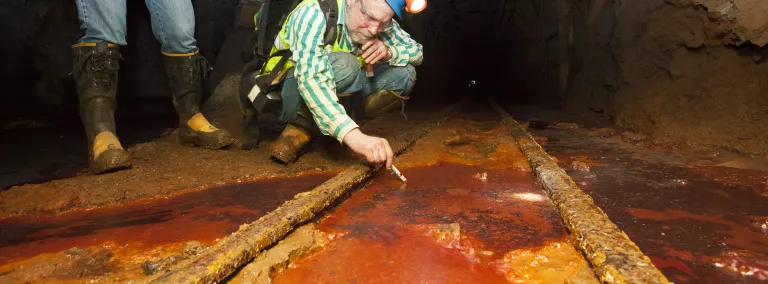 Dr. Dave Bergmann takes a sample from an underground area at SURF in between two train rails in rust colored fluid