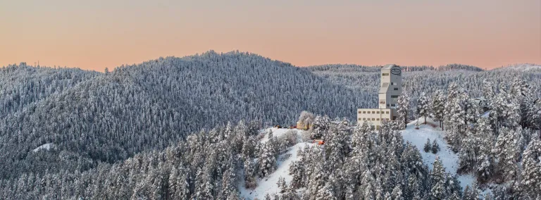This image shows the Ross Headframe at SURF surrounded by the snow covered Ponderosa Pines of the Black Hills on a cold morning as the sunrise colors the horizon in hues of red and orange.  