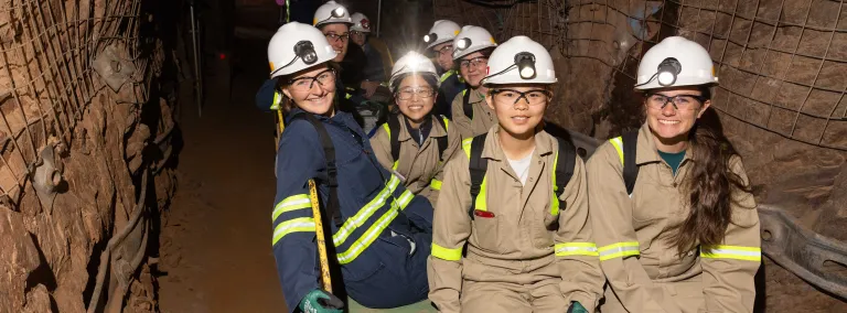 2024 cohort of Davis-Bahcall scholars on a tour of the underground at SURF.  This group is in full PPE sitting on a trolly in a drift underground.  They are all smiling. 