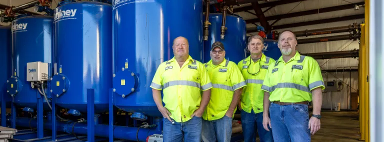 The Wastewater Treatment Plant crew at SURF posing in front of large blue settling tanks inside the WWTP:Pat Hasson, Cody Jones, Doug Coolley, and Dave Johnson.