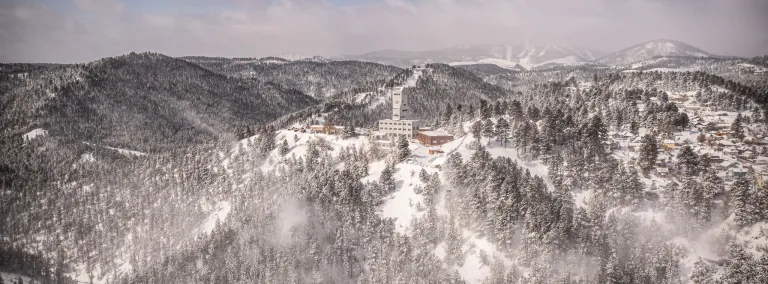 An aerial photo of SURF Ross Headframe in the winter covered in snow with the hills surrounding and Terry Peak Ski area in the background. 
