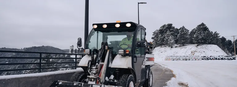 person drives a bobcat with shovel bucket on the front to remove snow from the sidewalk 