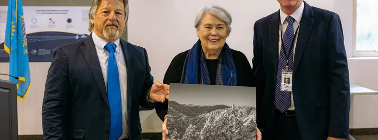 Three people stand facing the camera, the person in the middle holds a photo 