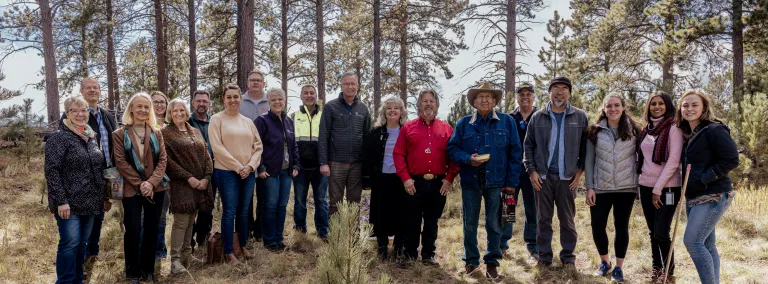 A group of people stand together outside with pine trees in the background 