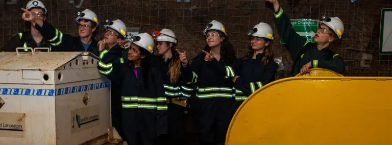 students in underground personal protective equipment stand in a drift and point into the air