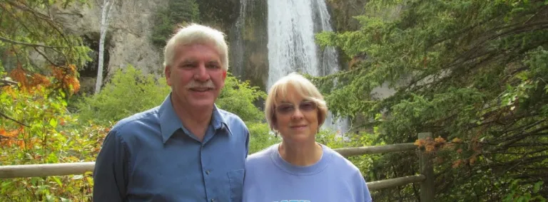 A couple stands in front of a waterfall surrounded by greenery