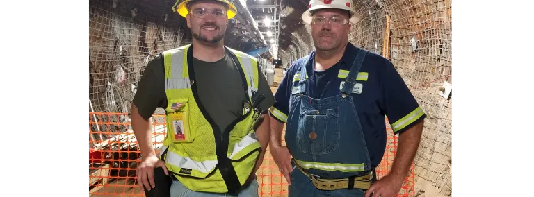 Two men in reflective gear and hard hats stand in a well-lit underground drift. 