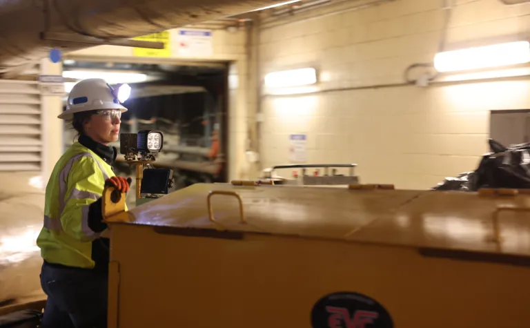 Sarah Wortman, an underground facilities technician at SURF, dressed in a hardhat, light, and reflective vest with red gloved, operates a locomotive on the 4850 Level undergroudn at SURF.