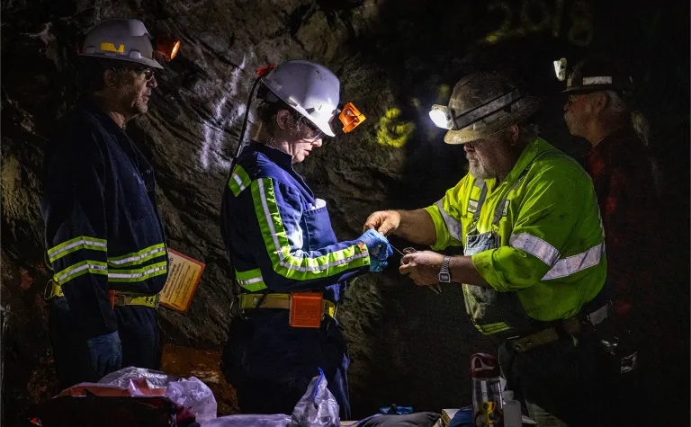 Two former Homestake workers, Al Pfarr (right) and Bryan Silvernail working with scientists Magdalena Osburn and Bradley Stevenson with the Deep Mine Microbial Observatory (DeMMO) to repair a biologic sampling site on the 800 Level of SURF. The photos is in a dark area, surrounded by rock inside the mine, individuals are wearing PPE and bright reflective clothing used underground.