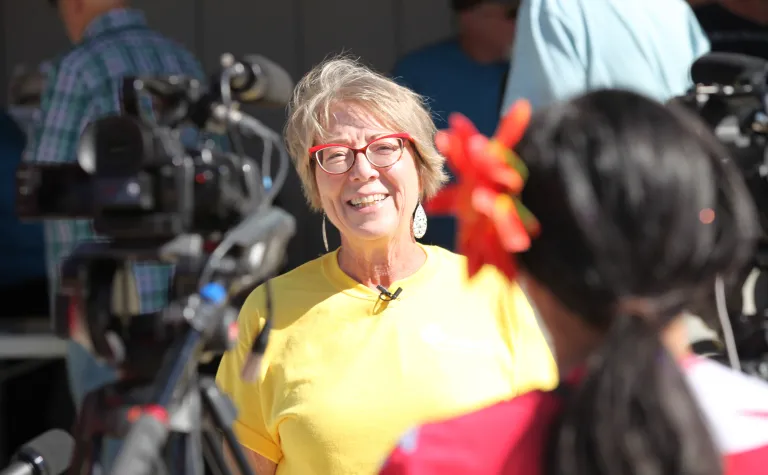 Constance Walter, wearing a bright yellow shirt and red glasses, smiles during TV interviews through a crowd of people.