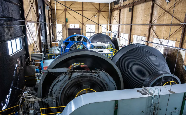 A shot of the ross hoists from the tower inside the hoist room, both hoists and their tapered double drum spools are seen form the side in a sun lit room through old windows.