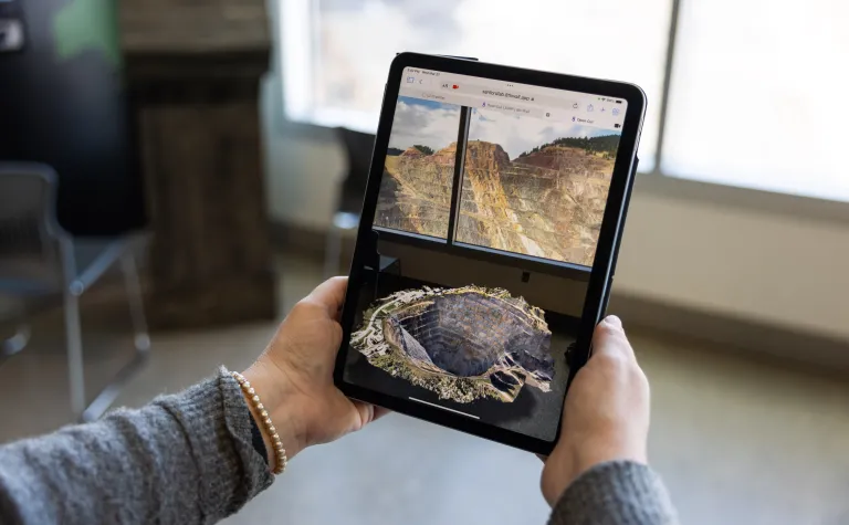 The view of the Open Cut in augmented reality at the Sanford Lab Homestake Visitor Center.  The viewer is holding a tablet screen with a 3D image of the Open Cut while looking out the window at the Open Cut.