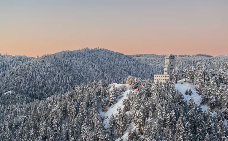 This image shows the Ross Headframe at SURF surrounded by the snow covered Ponderosa Pines of the Black Hills on a cold morning as the sunrise colors the horizon in hues of red and orange.  