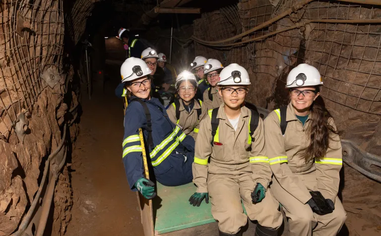 2024 cohort of Davis-Bahcall scholars on a tour of the underground at SURF.  This group is in full PPE sitting on a trolly in a drift underground.  They are all smiling. 