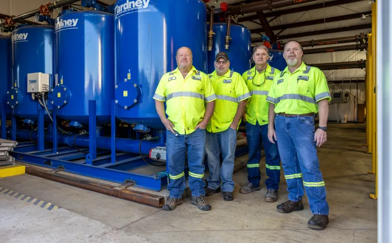 The Wastewater Treatment Plant crew at SURF posing in front of large blue settling tanks inside the WWTP:Pat Hasson, Cody Jones, Doug Coolley, and Dave Johnson.