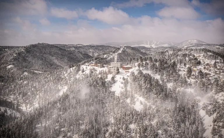 An aerial photo of SURF Ross Headframe in the winter covered in snow with the hills surrounding and Terry Peak Ski area in the background. 