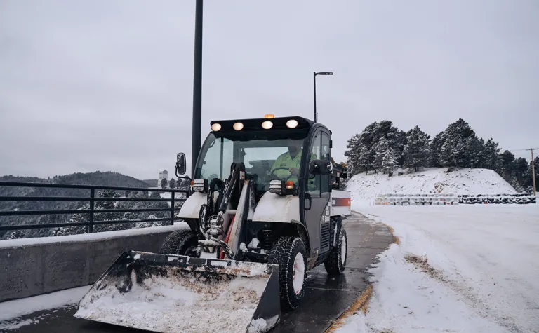 person drives a bobcat with shovel bucket on the front to remove snow from the sidewalk 