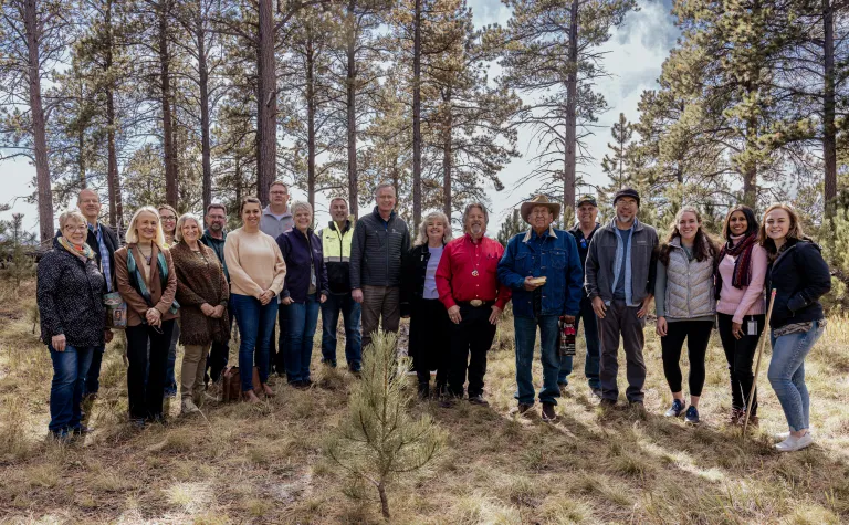 A group of people stand together outside with pine trees in the background 