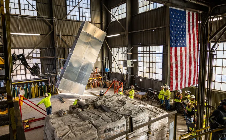 An APA is being lifted in the Ross Headframe with several infrastructure technicians standing by. A USA flag is hanging from the ceiling in the left side of the photo. 