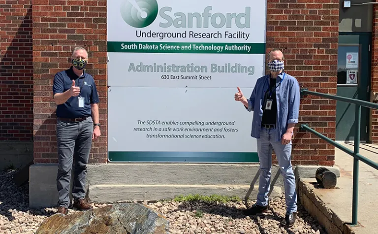 Two men in facemasks give thumbs up in front of the Sanford Lab sign