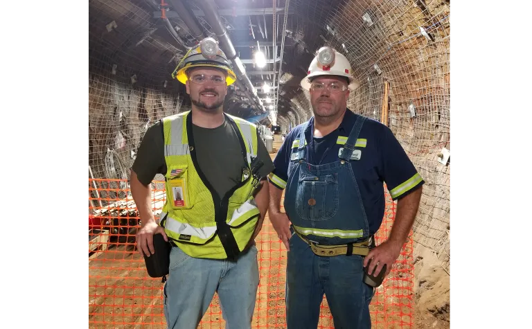 Two men in reflective gear and hard hats stand in a well-lit underground drift. 