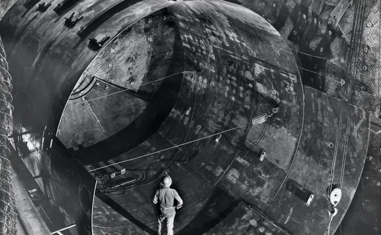 In a black and white photo, a man stands in a large, underground tank. 