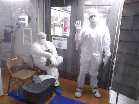 Two researchers in cleanroom garb, one os waving, the other sitting. 
