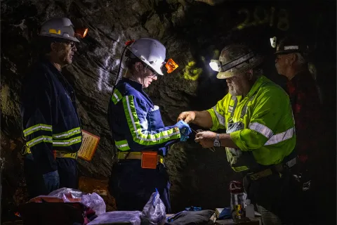 Two former Homestake workers, Al Pfarr (right) and Bryan Silvernail working with scientists Magdalena Osburn and Bradley Stevenson with the Deep Mine Microbial Observatory (DeMMO) to repair a biologic sampling site on the 800 Level of SURF.  The photos is in a dark area, surrounded by rock inside the mine, individuals are wearing PPE and bright reflective clothing used underground. 