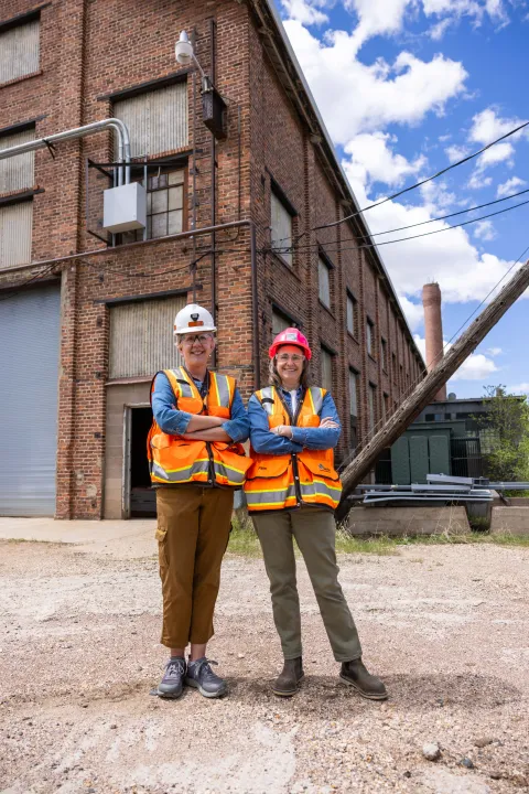 Bonita Goode (left), the environmental manager at SURF, stands next to Pam Hamilton, project manager at SURF, stand in full PPE in front of the Foundry Building, a brick structure on a spring day.