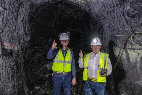 MIke Headley and Casey Peterson stand in front of a pile of rock underground wearing hard hats, lights, and reflective vests.