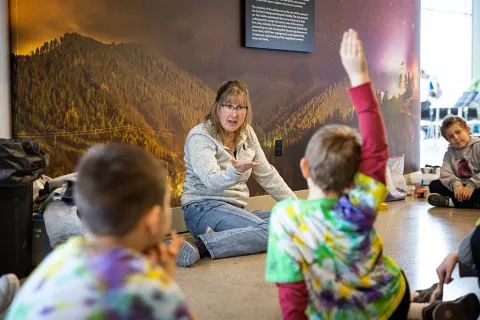 a teacher, Julie Dahl, instructus and interacts with young students while sitting on the floor of the Sanford Lab Homstake Visitor Center.  