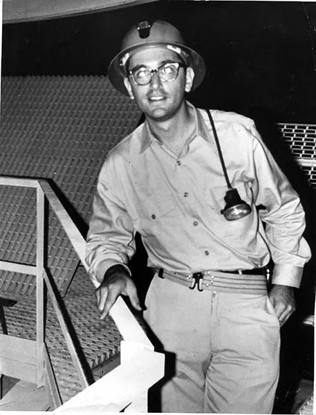 John Bahcall as a young man at the Homestake Mine wearing a hardhat, with a headlamp, in a black and white photo.