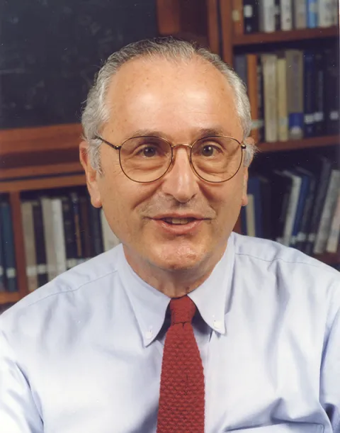 A photo of John Bahcall wearing a tie in front of a bookshelf. 