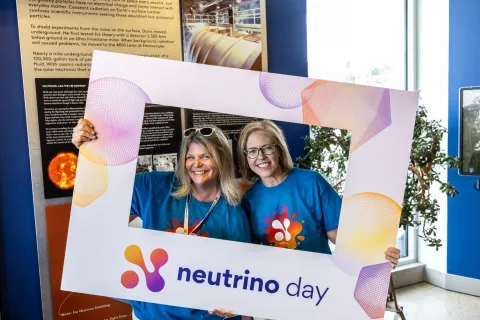 Two people smiling while holding the Neutrino Day selfie frame. 