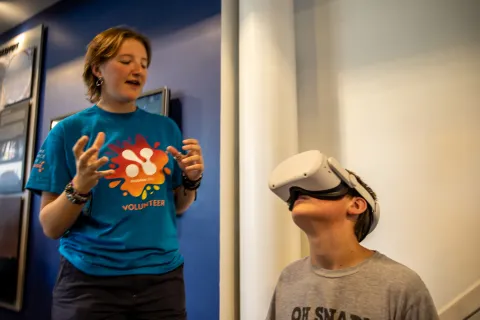 A child looks though VR goggles while a volunteer explains what they are seeing. 