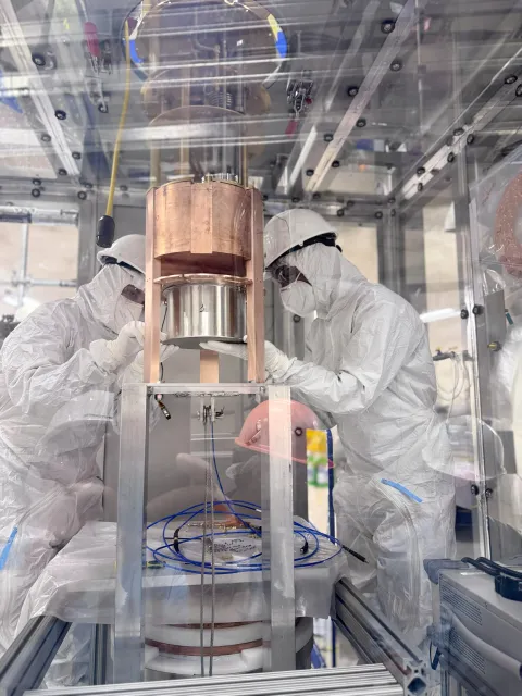 Two researchers in cleanroom garb install parts inside a laboratory. SNOLAB’s Cryogenic Underground Test Facility (CUTE) cleanroom, installing the test qubits into the CUTE dilution fridge.
