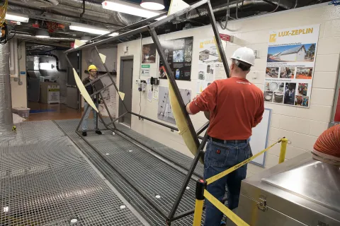 Two people stand at either end of a metal frame box with a rotary mechanism. 