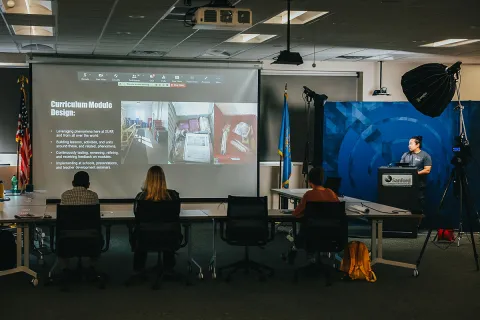 a person stands in front of a powerpoint, giving a presentation to a room of other people 
