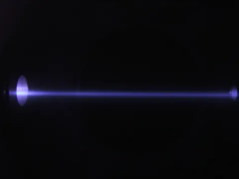 Photo of an ion beam passing through the gas of a gas target. It is an older photograph from 2011, thus, not from CASPAR, but the setup is the same. The beam enters through the aperture (not so much a pinhole though) from the left, travels through the gas and excites the atoms of the gas, which emit light during de-excitation.